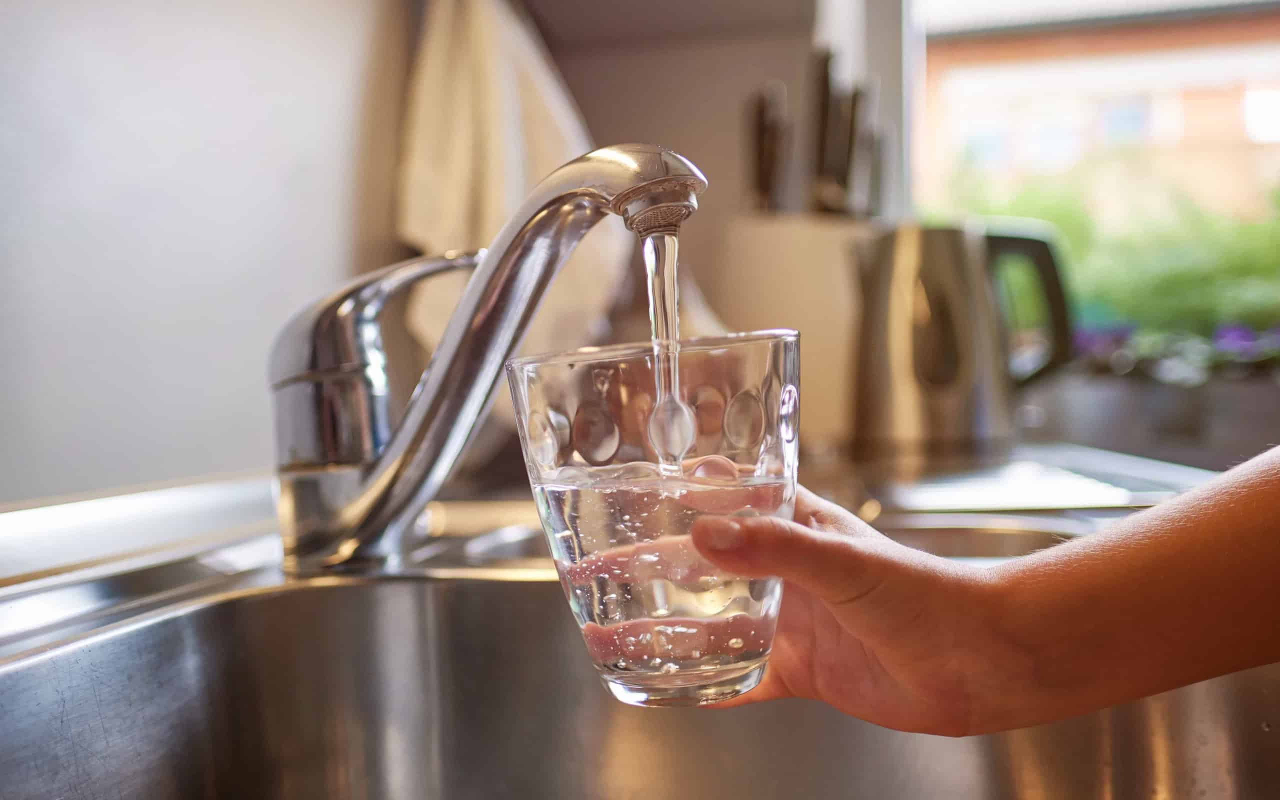 Eine Hand hält ein Glas unter den Wasserhahn in einer Küche und befült es mit Leitungswasser.