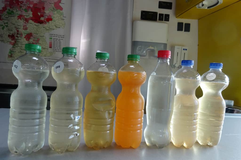 Sieben Brunnenwasserproben abgefüllt in Flaschen stehen auf dem Tisch. Die Proben sind teilweise mit Eisen belastet.